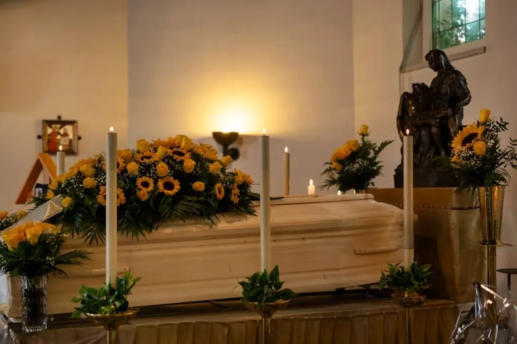 Cercueil en bois clair orné de fleurs jaunes et cierges pour cérémonie funéraire.